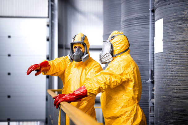 Factory worker standing by large metal storage tanks with acids wearing yellow protection suit, gas mask and gloves explaining trainee process of chemicals production inside the plant. - Photo, Image