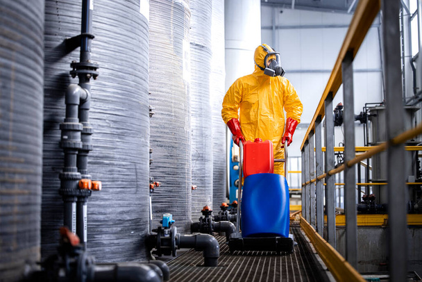 Factory worker walking by storage acid tanks and pushing cart with chemicals inside production plant. - Foto, Imagen