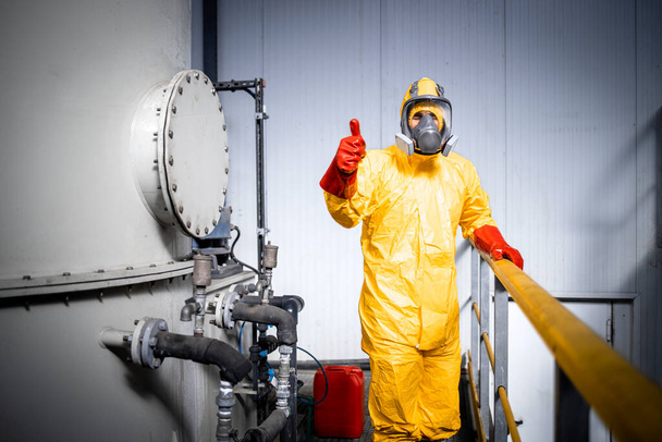 Portrait of factory worker in protective yellow suit, gas mask and gloves holding thumbs up inside chemicals production plant. - Foto, imagen