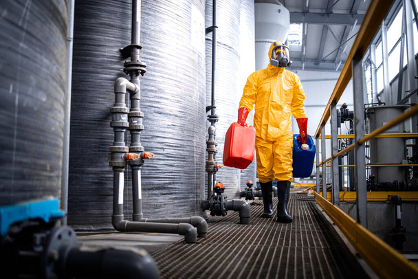 Factory worker in protective equipment walking by acid storage tanks and carrying aggressive and dangerous chemicals. - Foto, imagen