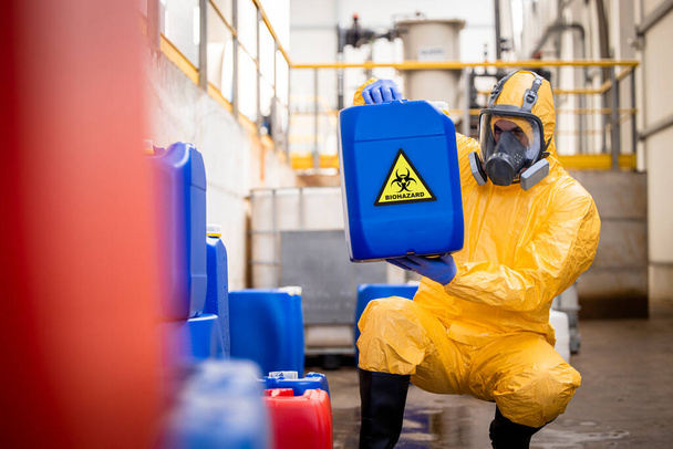 Factory worker in protective equipment and gas mask holding plastic can with toxic chemicals and biohazard waste inside recycling plant. - Fotó, kép