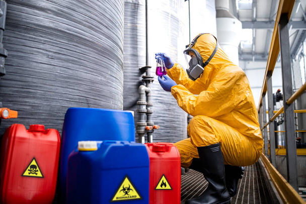 Chemical factory worker wearing protection suit and gas mask testing quality of chemicals. In background storage tanks for acid and industrial interior. - Foto, Imagen