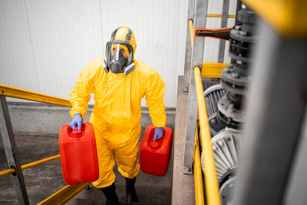 Trabajador totalmente protegido en traje de materiales peligrosos y máscara de gas que transportan productos químicos. Interior industrial. - Foto, imagen