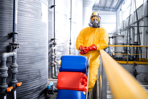 Portret van fabrieksarbeider in beschermingspak en gasmasker die kar met chemicaliën duwt door grote zuuropslagtanks in productie-installatie. - Foto, afbeelding