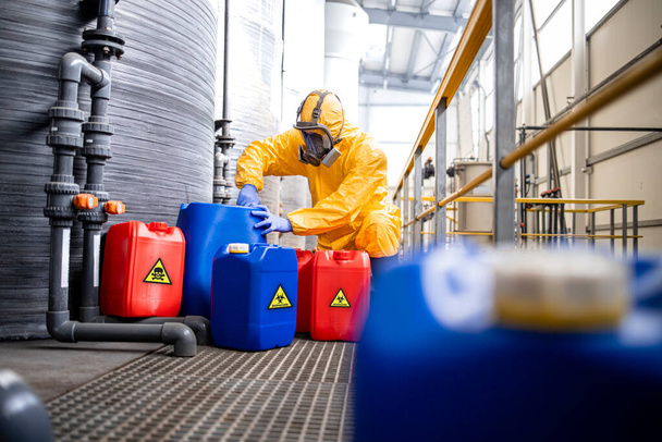 Factory worker in hazmat suit and gas mask filling plastic container with chemical and hazardous waste. Chemicals factory interior with acid storage tanks. - Zdjęcie, obraz