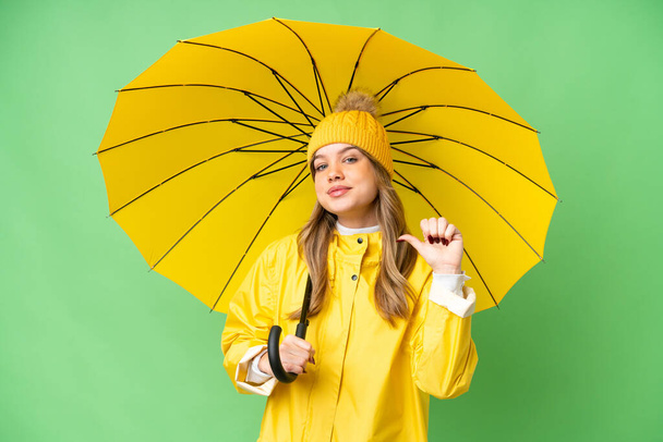 Νεαρό κορίτσι με αδιάβροχο παλτό και ομπρέλα πάνω από απομονωμένο υπόβαθρο κλειδί chroma υπερήφανος και αυτάρεσκος - Φωτογραφία, εικόνα