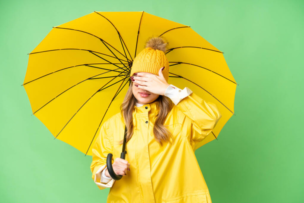 Молодая девушка в непромокаемом пальто и зонтике на изолированном хроматическом фоне закрывает глаза руками. Не хочу ничего видеть. - Фото, изображение