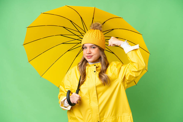 Yağmura dayanıklı ceketi ve şemsiyesi olan, izole edilmiş krom anahtar geçmişi olan genç bir kız güçlü bir jest yapıyor. - Fotoğraf, Görsel