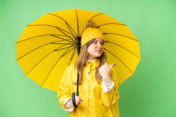 Menina com capa impermeável e guarda-chuva sobre fundo chave croma isolado apontando para o lado para apresentar um produto - Foto, Imagem