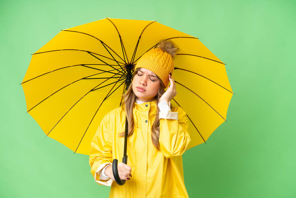 Chica joven con capa impermeable y paraguas sobre fondo de croma clave aislado con dolor de cabeza - Foto, Imagen