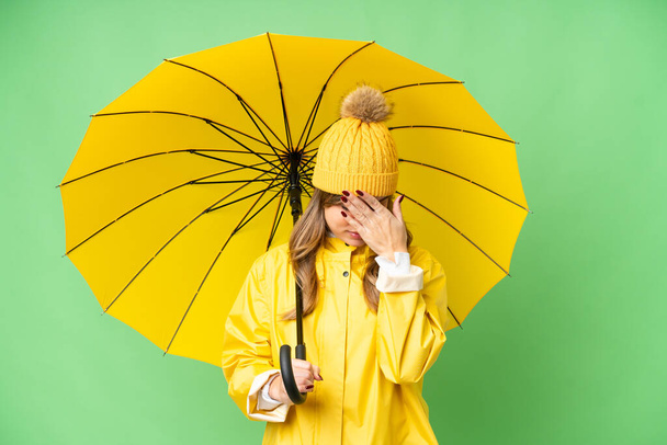 Jeune fille avec manteau imperméable et parapluie sur fond isolé chroma clé avec expression fatiguée et malade - Photo, image