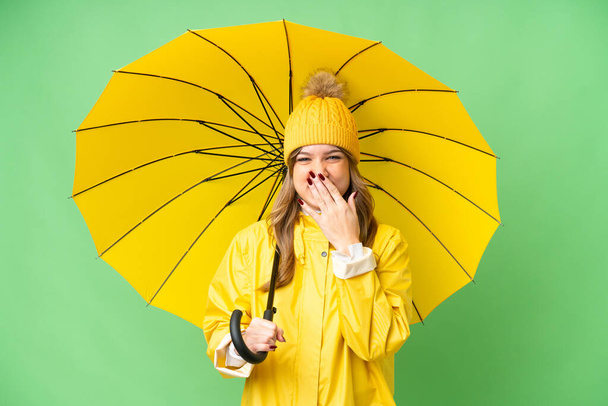 Yağmura dayanıklı ceketi ve şemsiyesi olan genç bir kız izole edilmiş krom anahtar arka planda mutlu ve ağzı elle kaplanmış gülümsüyor. - Fotoğraf, Görsel