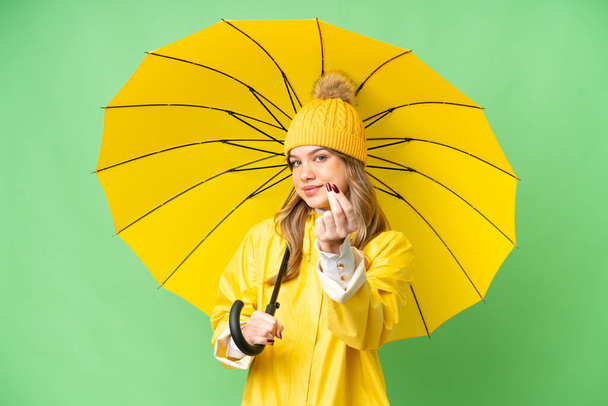 Menina com capa impermeável e guarda-chuva sobre fundo chave chroma isolado fazendo gesto de dinheiro - Foto, Imagem
