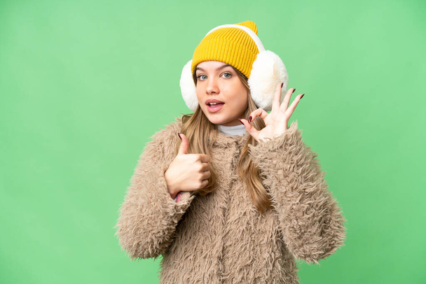 Chica joven con muffs de invierno sobre fondo de croma clave aislado que muestra signo de ok y el pulgar hacia arriba gesto - Foto, Imagen