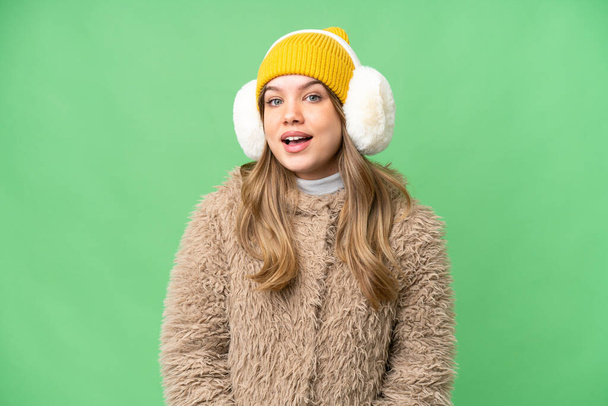Chica joven con muffs de invierno sobre fondo de croma clave aislado con expresión facial sorpresa - Foto, imagen