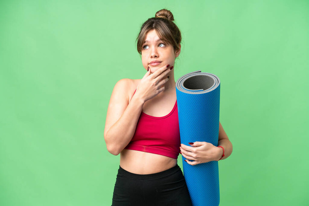 Junges Sportmädchen geht zu Yoga-Kursen, während sie eine Matte über isolierten Chroma-Schlüsselhintergrund hält und Zweifel hat - Foto, Bild