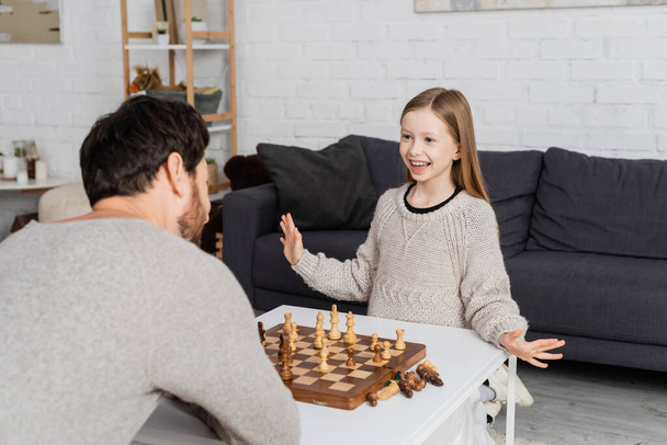 cheerful preteen girl showing wow gesture near father and chessboard on coffee table - Foto, imagen