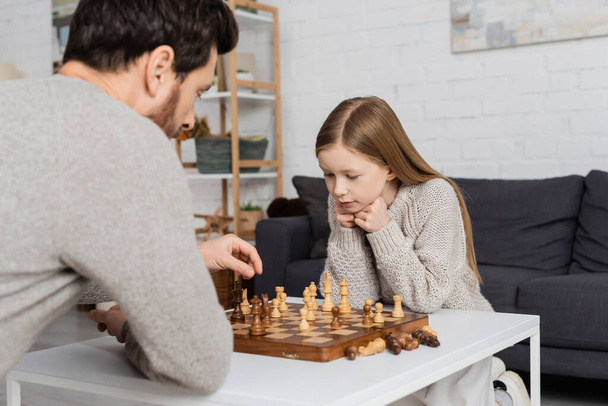 preteen girl thinking near dad and chessboard on coffee table in living room - Photo, Image