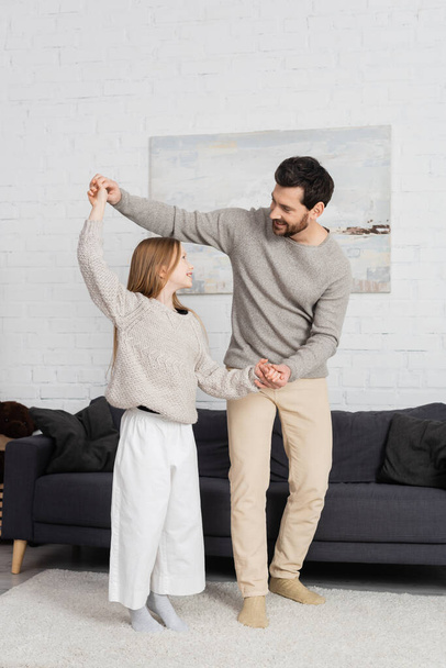 full length of positive bearded man teaching daughter to dance at home - Foto, afbeelding