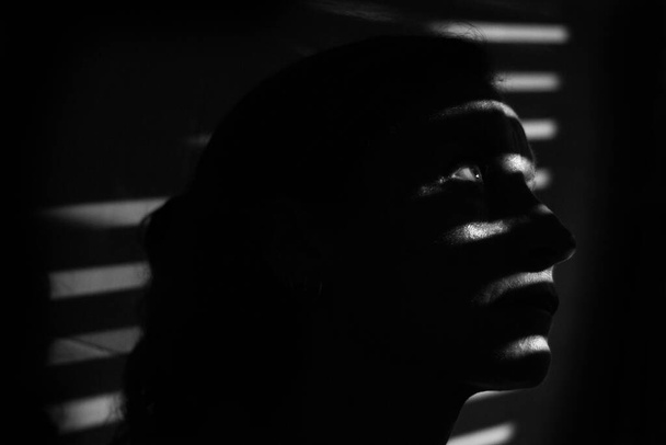 Yüzünde çizgilerin gölgesi olan karanlıkta genç bir kadının siyah beyaz portresi. Gösterişsiz model oturumu konsepti - Fotoğraf, Görsel