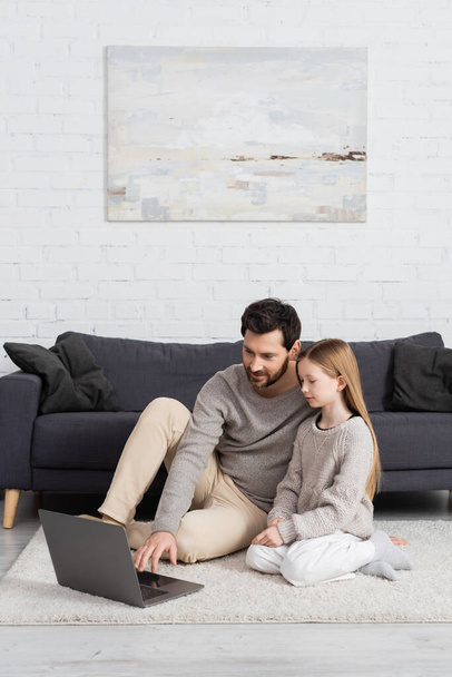 preteen girl sitting near father using laptop on floor carpet in living room - Foto, immagini