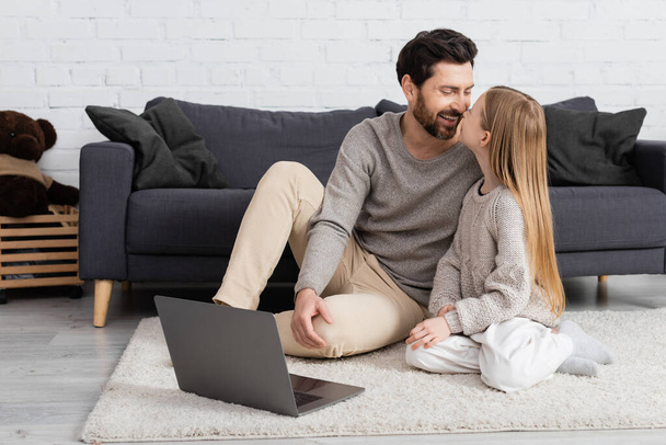 cheerful father and daughter smiling at each other near laptop on floor in living room - 写真・画像