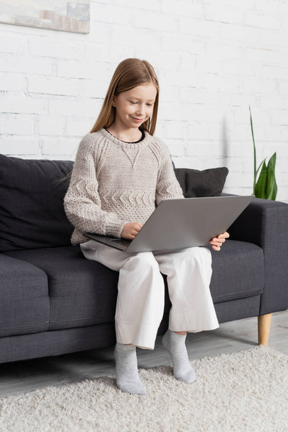 full length of smiling girl using laptop while sitting on couch in living room - Foto, imagen