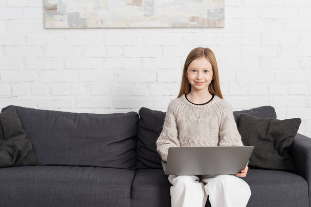 pleased girl sitting on couch with laptop and smiling at camera - Photo, image