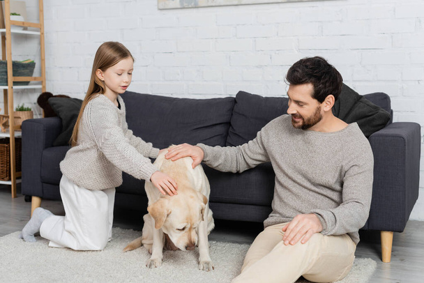 happy father and daughter sitting on floor near couch and petting labrador dog - Foto, Imagem