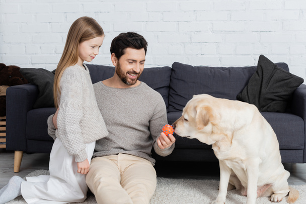 fröhlicher bärtiger Mann umarmt Tochter, während er Spielzeug in der Nähe von Labrador-Hund im Wohnzimmer hält - Foto, Bild