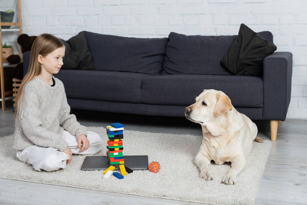 chica feliz y perro labrador mirándose unos a otros cerca de bloques de madera juego y portátil en el suelo en la sala de estar - Foto, Imagen