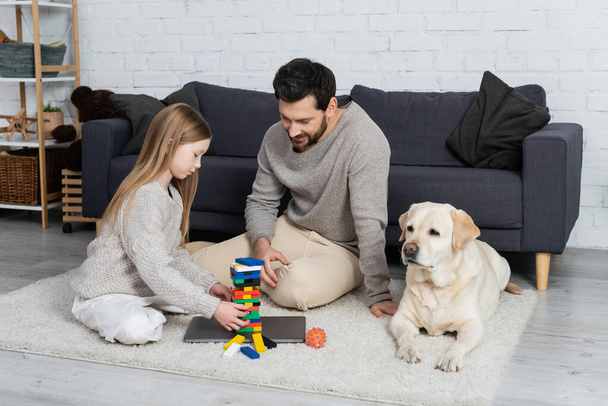 gelukkig bebaarde man met tiener dochter spelen houten blokken spel in de buurt van laptop en labrador hond op de vloer thuis - Foto, afbeelding