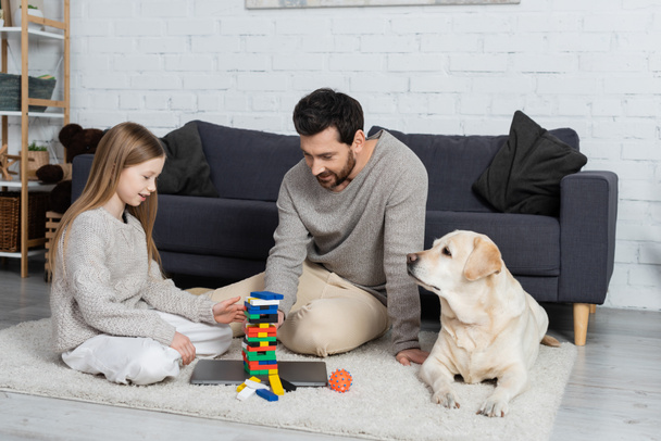 smiling father and daughter playing wood blocks game near labrador dog on floor in living room - Photo, Image