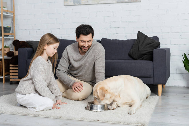 labrador dog drinking water from bowl near cheerful father and daughter on floor carpet at home - Foto, Imagem