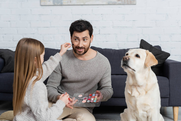 niña preadolescente sosteniendo paleta de maquillaje mientras juega con papá sonriente cerca del perro labrador en la sala de estar - Foto, imagen