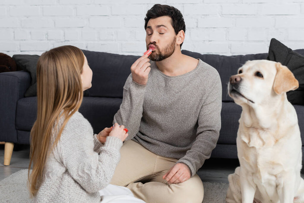 bärtiger Mann mit geschlossenen Augen, der Lipgloss aufträgt, während er mit Tochter in der Nähe von Labrador-Hund im Wohnzimmer spielt - Foto, Bild
