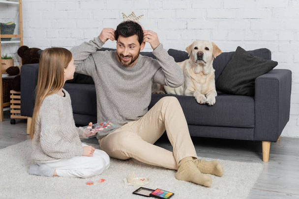 cheerful bearded man wearing toy crown while sitting on floor near daughter and dog lying on sofa - Foto, immagini