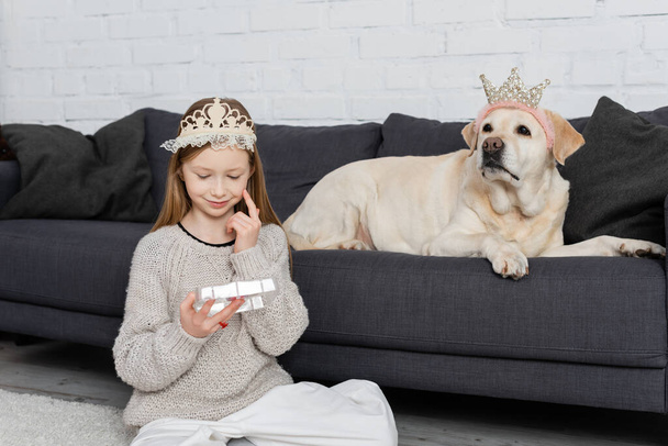 menina pré-adolescente feliz na coroa olhando para a paleta de maquiagem e sentado no tapete perto labrador  - Foto, Imagem