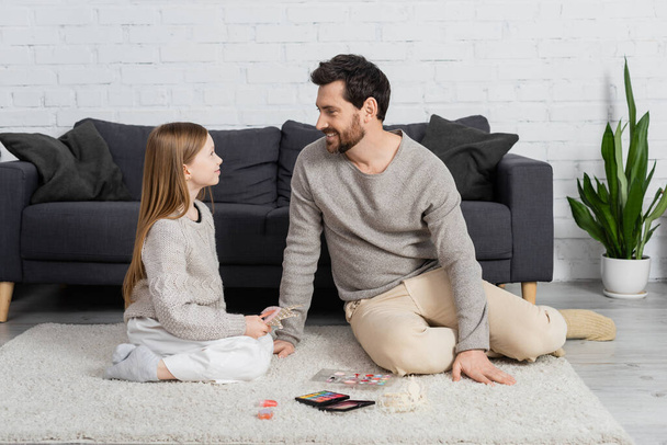 szczęśliwy ojciec patrząc na uśmiechniętą córkę siedząc na dywanie w pobliżu kosmetyków dekoracyjnych  - Zdjęcie, obraz