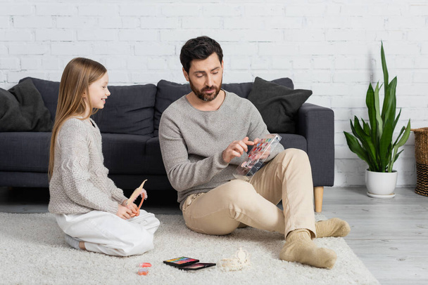 bearded father pointing at makeup palette while sitting on carpet with happy daughter  - Foto, Bild