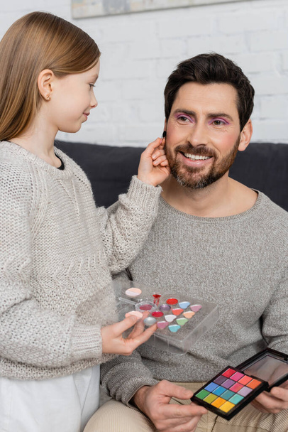 preteen girl holding cosmetic brush while applying beauty product on cheek of bearded father  - Φωτογραφία, εικόνα