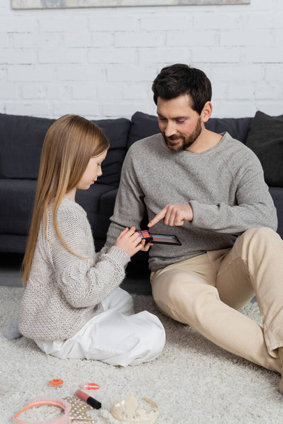 happy father pointing at eye shadow palette near daughter in living room  - Photo, Image