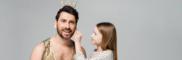 happy kid applying decorative cosmetics on face of bearded father in crown and dress isolated on grey, banner  - Foto, Bild