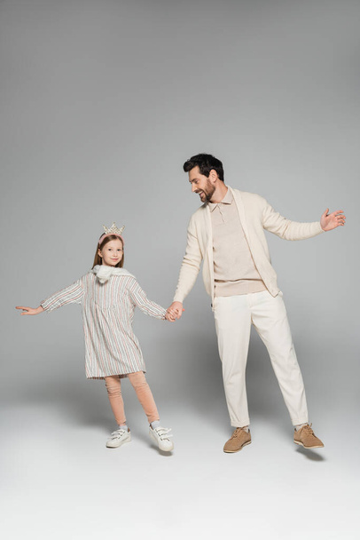 full length of cheerful father and daughter in dress holding hands on grey  - Foto, afbeelding