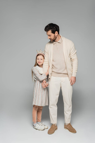 full length of happy girl in dress and crown hugging father in white outfit on grey  - Photo, Image