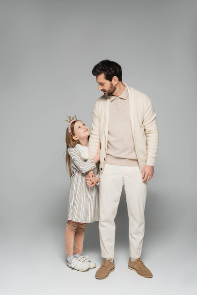 full length of cheerful girl in dress and crown hugging father in white outfit on grey  - Photo, image