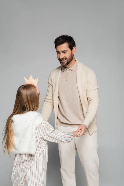 happy bearded man looking at daughter in crown while holding hands on grey  - Fotó, kép