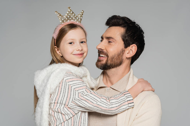 fröhliches Kind in Krone blickt in die Kamera, während es Vater isoliert auf grau umarmt  - Foto, Bild