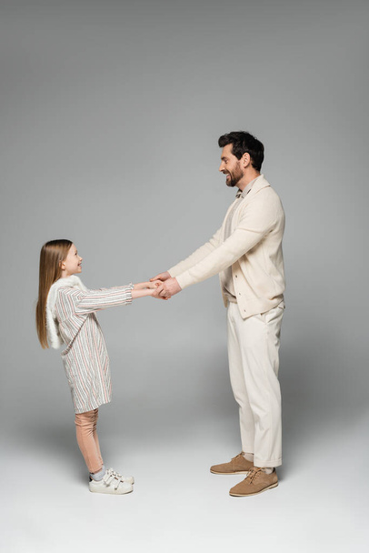 side view of happy father and daughter holding hands while looking at each other on grey  - Zdjęcie, obraz