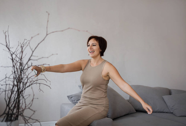 happy, contented beautiful woman over 30 in a tight dress, happily spreading her arms, smiling, sits on the couch. elegant business lady celebrating triumph, joy from good news. Positive thinking - Photo, Image
